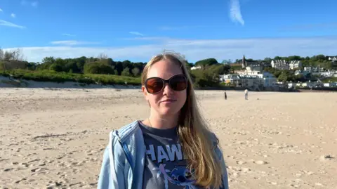 Karen Taylor wears sunglasses, a light blue hoodie, and a dark T-shirt with a cartoon character and the word “Stitch” stands on a wide sandy beach. Low grassy dunes stretch behind her, with several people walking in the distance and a cluster of light-coloured buildings and trees along the shoreline. The sky is bright blue with scattered clouds.