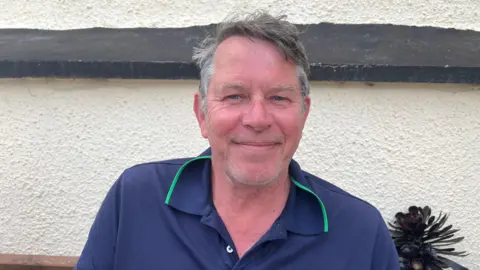 A man smiles at the camera with a blue t shirt with green collar