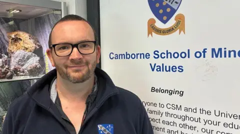 Matt Tonkin wearing a dark coloured fleece with a Camborne School of Mines logo on it and standing in front of a Camborne School of Mines sign to the right with a display of geological rocks to the left.