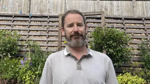 Jon Dean A man with short dark hair and a short dark beard is wearing a grey top and standing in front of a garden fence