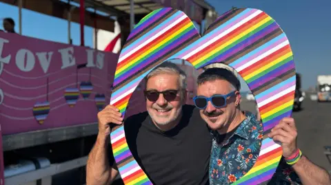 Two men stand within a cut-out rainbow heart