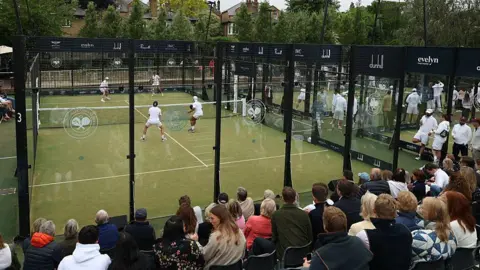 Getty Images A view of a padel court during a tournament. Spectators sit in stands around the court. The court is separated from the crowd by a glass box. Two players play on either side of the court dresses in white sports clothes. 