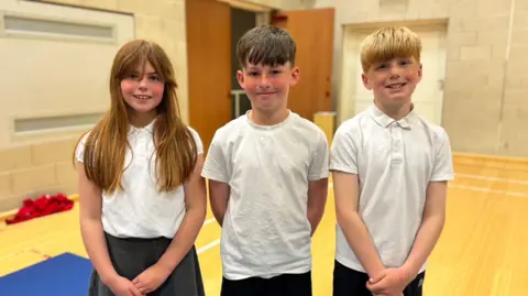 Lilly (left), Teddy (middle), and Freddie (right) are standing in a sports halls, all wearing white t-shirts and black bottoms. Lilly has long brown hair and a fringe, Teddy has short dark hair and Freddie has blonde/strawberry blonde hair. They are all smiling.