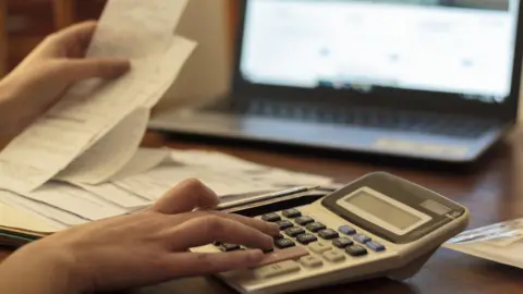 Getty Images An anonymous person sits at a desk with a pile of receipts in one hand and the other hovering over a calculator. An open laptop is visible in the background.