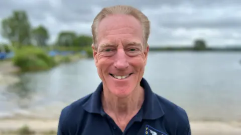 An older man with blondy grey hair smiles as he stands in front of a lake on a cloudy day. He is wearing a navy polo shirt.