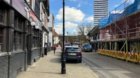 Street view of Spon Street in Coventry. There is scaffolding and yellow barriers on the right and a row of shops and restaurants on the left. There are parked cars in the background and a black lamppost in the middle of the photo.