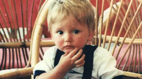 A picture of a little boy with blonde hair wearing a white shirt, sat on a rattan chair