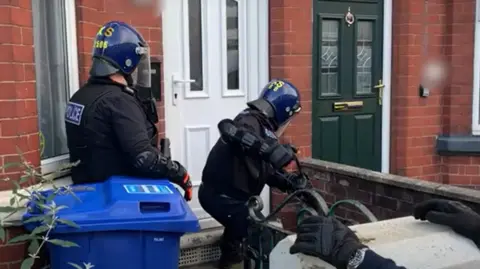 South Yorkshire Police A police raid in progress at a residential property. Three officers, dressed in black tactical uniforms and helmets marked "POLICE" and "TSG" (Tactical Support Group), are actively engaged in forced entry. One officer is using a battering ram to break down a white front door of a red-brick house. Another officer stands behind him, holding an orange tool, possibly used for breaching or support. A third officer is partially visible on the right side, wearing gloves and assisting in the operation.
Next to the door being breached, there is a blue recycling bin. In the background, a green door with a brass mail slot and knocker can be seen.