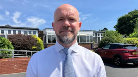 Dave is stood in the school ground with buildings behind him and looking directly at the camera. He has a white shirt on and blue tie. He has a beard.
