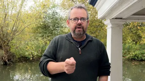 David Thomas being interviewed on the Corpus Christi Barge. He is looking at the interviewed and away from the camera. He has his right rang in a fist, aiming to make a point. The river is behind him. It is cloudy.