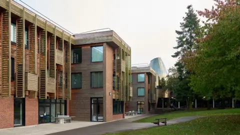 St Regis CE Academy Large modern-looking buildings with two large trees and grass on the right of the picture