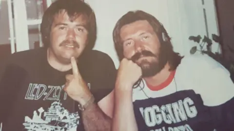 Allan Weaver Two men sat next to each other. Both have dark hair and the man on the left has a moustache and the man on the right has a beard. Both had t-shirts on. The t-shirt worn by the man on the left is a Led Zeppelin t-shirt and has some of the mand's name visible. 