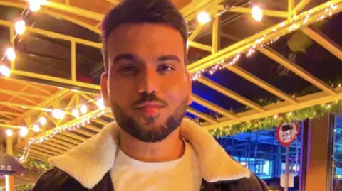 Mohammed Armaan A person is standing under a covered outdoor structure decorated with rows of warm string lights. The roof has a yellow frame with a wooden finish, and the background includes greenery and a few visible signs. The individual is wearing a white crew-neck shirt layered under a dark jacket with a light-colored shearling collar. The setting appears festive or part of an outdoor dining area.