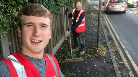 Matthew Locke with short brown hair wears an orange hi-vis and grey t-shirt with another man behind him wearing an orange hi-vis sweeping leaves up off the pavement. They are smiling.
