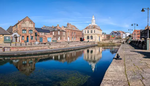 KLWNBC View of King's Lynn Quayside showing a body of water with red brick house either side. Custom House is in the background, a white stone imposing building. The photograph is taken on a sunny, cloudless day and Custom House's reflection can be seen in the water. 
