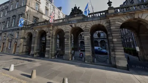The front of Edinburgh City Chambers has stone pillars with flags hanging from it. Through the archways leads to a courtyard and the entrance to the City Chambers.