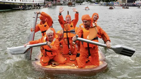 Colin Moody Six people dressed in bright orange overalls, paddling across Bristol's harbourside on a raft, using large oars which have been designed to look like knives and forks. The raft they are floating on looks like a piece of toast, and they are meant to resemble beans.