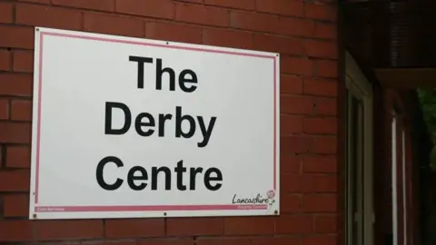 A white sign on a red brick wall, with the black writing saying, The Derby Day Centre, and Lancashire county council in the bottom right hand corner