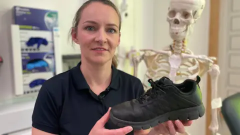 A woman with her hair tied back holds up a black trainer. She is standing in a medical room with a life-size skeleton