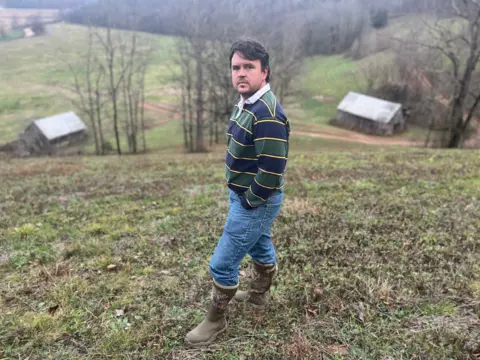 BBC/Ellie House Seorang pria di atas bukit melihat ke arah kamera, mengenakan jeans dan sepatu bot, ada lumbung dan perbukitan di kejauhan.