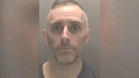 Police custody image of Scott Cameron. He has short grey hair and stubble wearing a brown t-shirt.