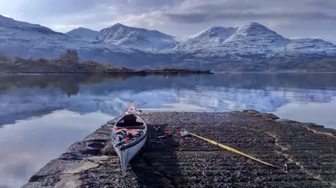 Colin Cowie A kayak and oar on a jetty in front of a loch, with snow-covered hills in the distance, reflected in the water.