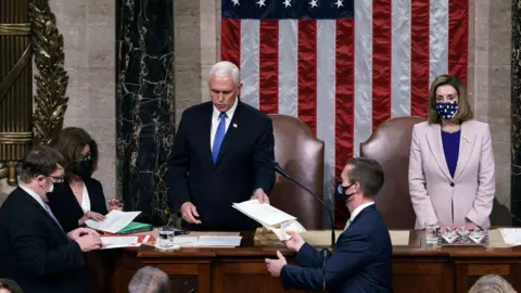 Getty Images Mike Pence stands in Congress and hands papers to staff as Speaker of the House Nancy Pelosi stands beside him in a mask