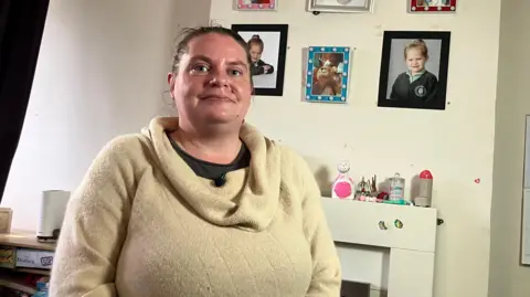 Maggie sitting in her living room at all. Her dark-brown hair is pulled back into a ponytail and she is wearing a cream-coloured jumper. Her children's school pictures are hanging on the wall in the background.