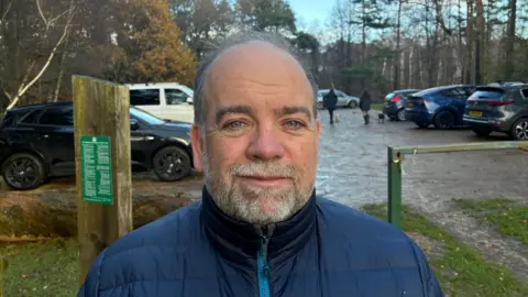 Chris McHugh/BBC Richard Burke of Forestry England poses for the camera in front of a muddy gravel car park which is surrounded on all sides by tall trees. Vehicles can be seen in the car park behind him parked in rows on either side.