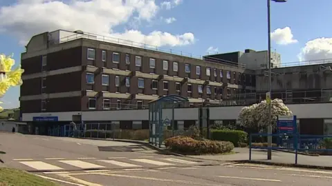 A view of the front of North Devon District Council. The building is brown and has four levels with many small windows. There is a sign to the right of the building and a zebra crossing at the centre.
