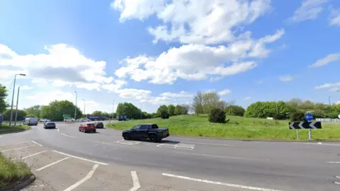 A street view image of a large roundabout with grass in the centre. The sky is blue and cloudy