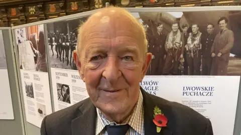 Jan Illaszewicz wearing a shirt, tie and blazer with a poppy attached to it stood in front of an exhibition of the 307 Squadron in Exeter