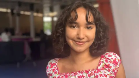 Maya Wijayasiri, who is wearing a pink floral top, is smiling at the camera. 