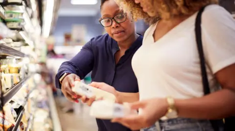 Getty Images Two women, one in a blue shirt and glasses, compare cheese in the aisle of a grocery store