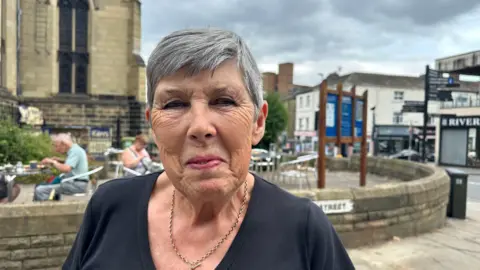 A woman with a grey pixie cut looks towards the camera, she is wearing a black v-neck t-shirt and a gold necklace. Behind her is a brick wall and people on silver metal outdoor seating eating and drinking.