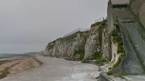 A Google Street image of a stretch of concrete promenade with a short, white chalk cliff behind it to the right, and the beach to the left.