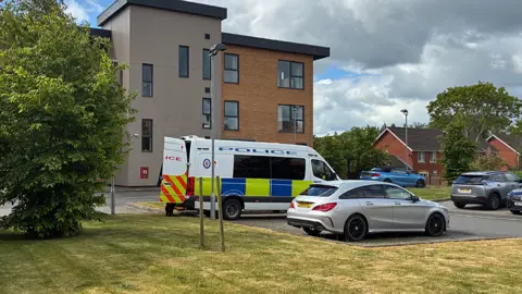 A police van in a car park, near four other vehicles. There is closely cropped grass in the foreground and a three-storey building in the background