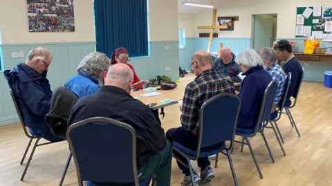 A group of people are sitting round a table praying and in the corner there is a cross against the wall.