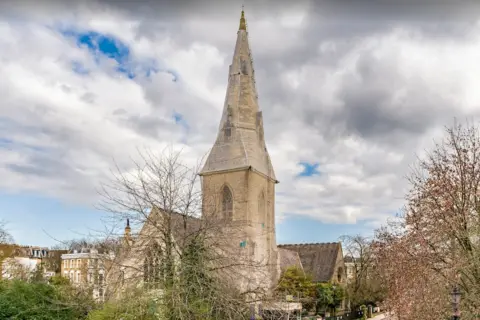 Historic England St Andrew's church spire rises above the treeline within a residential area. The spire is stone and has slightly weathered looking patches on it. 
