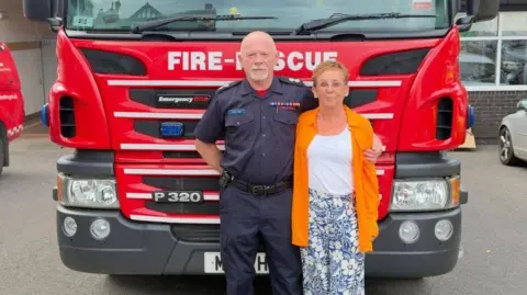 Shropshire Fire and Rescue Service A bald man with a navy firefighter uniform on, with his arm around a woman who has short orange hair and glasses. They are standing in front of a red fire engine