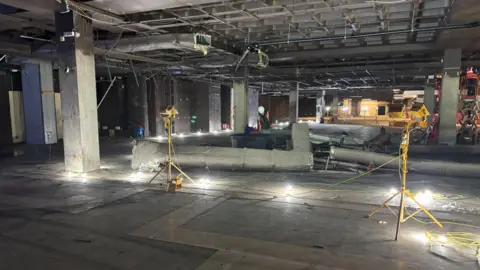 Jodie Halford/BBC A large, warehouse-like space is lit by small lamps on the floor. Spirit levels and other constructions equipment is dotted around the concrete floor.