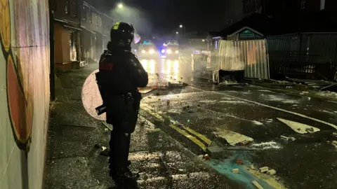 Riot police officer stood on street with bricks and other damage on ground