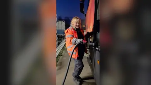 Woman with blonde hair and orange high vis jacket smiles while filling lorry with fuel.