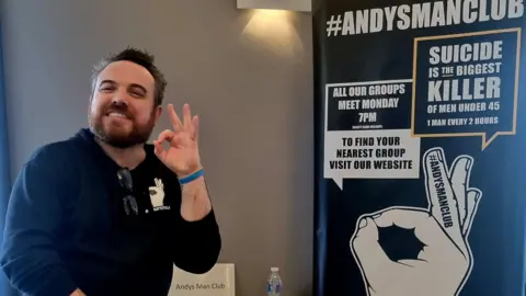 Steve Doyle, from Andysmanclub, is standing to the left holding up one hand and smiling. Next to him is a poster detailing a men's support group that meets every Monday.