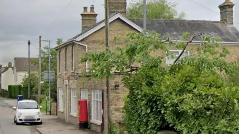 A street view of the old post office. It is a light coloured brick building with a post box outside. A small white car is parked outside and there is a large leafy green bush to the side.