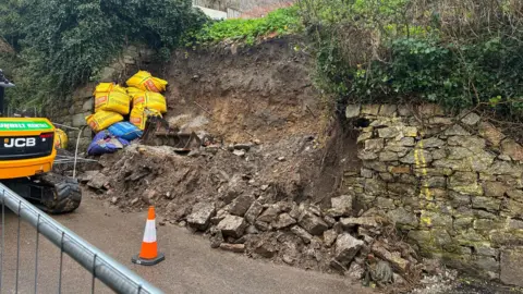 The collapsed wall. The stone has fallen to the base and exposed a pile of earth and sand. There is a safety barrier, a traffic cone, sacks of building materials and a mini-JCB next to the collapse.