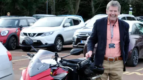 Gordon Castle Gordon Castle is standing next to a large motorbike wearing trousers, a pink shirt and a jacket with a lanyard around his neck with a name tag on it.