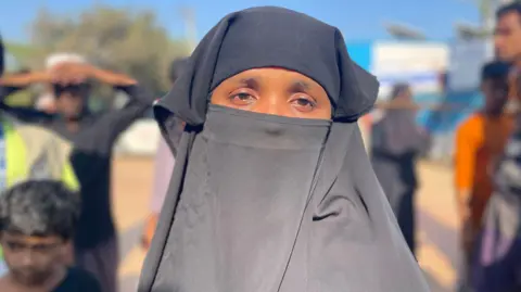 A woman wears a black loose costume, covers her face leaving her eyes