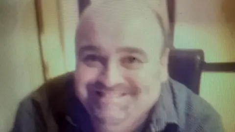 Close up image of Martin Fletcher. He is wearing a striped shirt and is sitting in a chair. He has dark stubble and dark eyebrows.