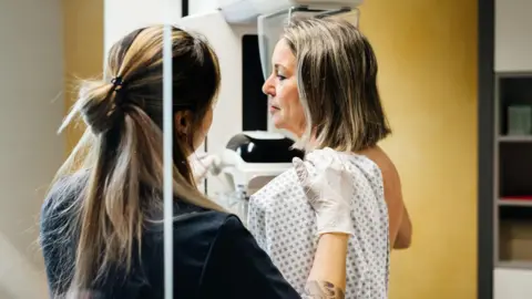 A nurse with brown hair half tied back is faced away and providing assistance to a woman with shoulder length hair in a hospital gown who is undergoing a mammogram in a modern medical facility. 